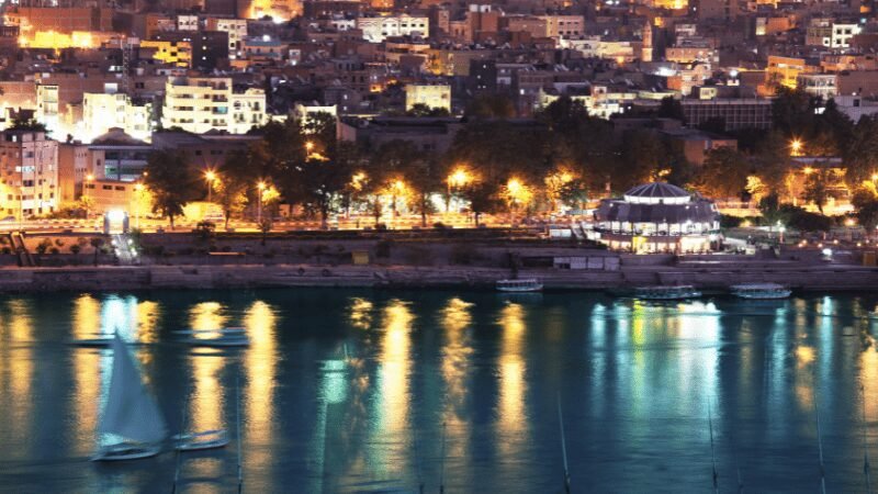 Evening view of a cityscape by the river, illuminated and reflected on the water's surface.
