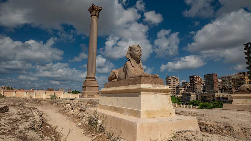 A lone Sphinx statue in front of a tall column amidst ruins, under a blue sky