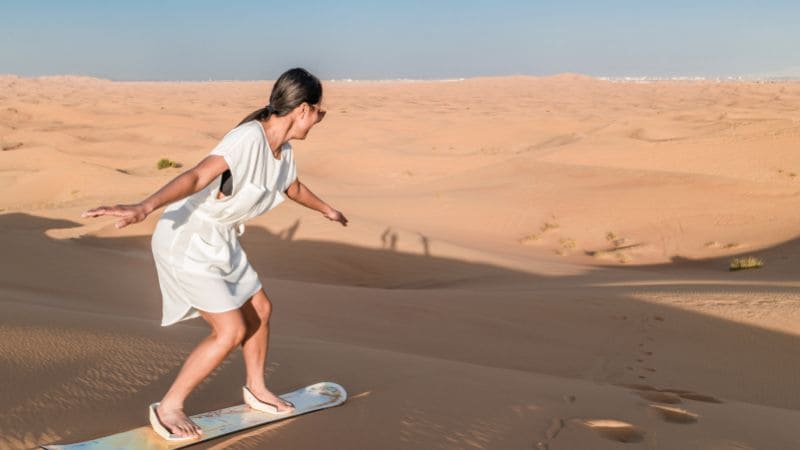 A person sandboarding down a desert dune, leaving a trail in the sand.