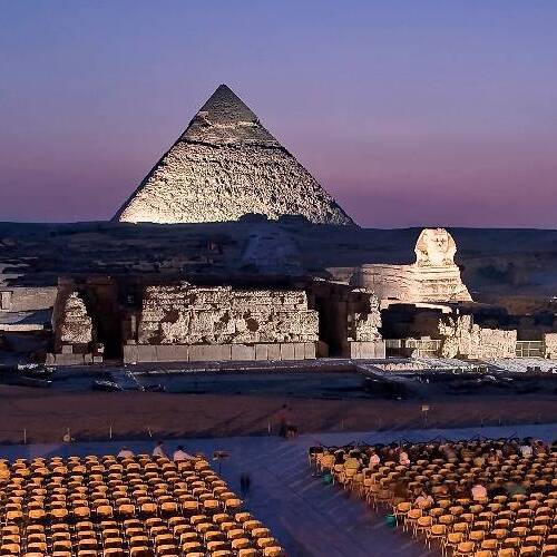 A view of the Great Pyramid and the Sphinx in Giza with rows of empty chairs in the foreground, under a pink twilight sky