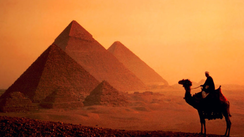 A silhouette of a camel rider in front of the Pyramids of Giza at dusk.