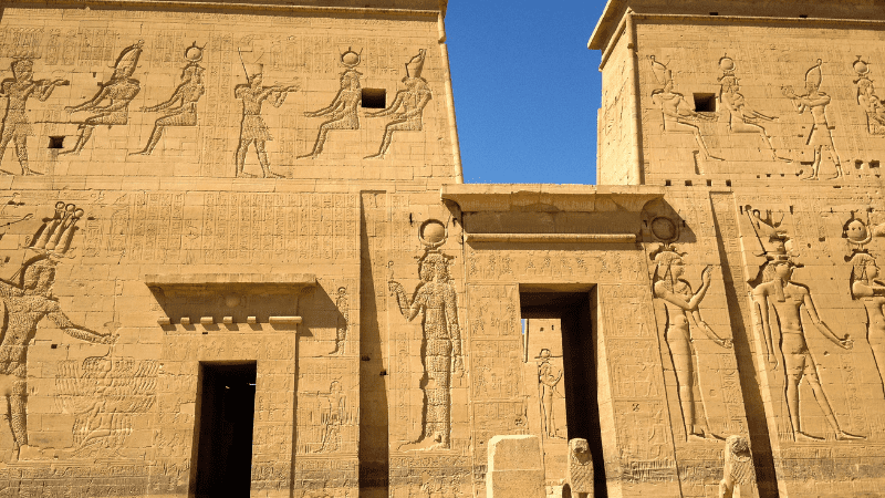 Ancient Egyptian temple facade with carved figures and hieroglyphs in bright sunlight.