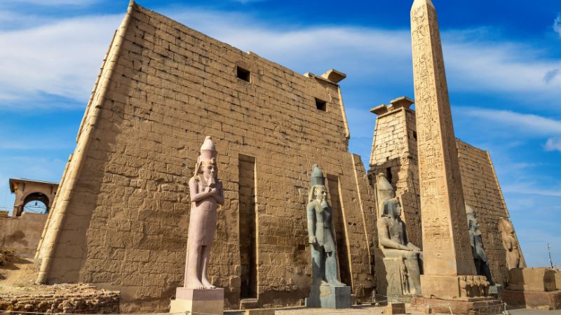 "Stone statues and obelisk at the entrance of Luxor Temple under a blue sky."