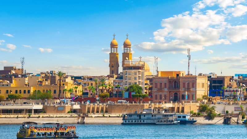 A riverside view with colorful buildings and prominent church spires under a blue sky.