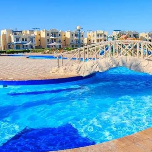 A vibrant swimming pool with clear blue water, a white stone bridge, and beige vacation apartments under a clear sky.