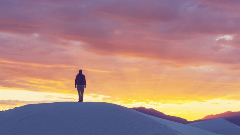 "Silhouette of a person standing atop a sand dune at sunset."