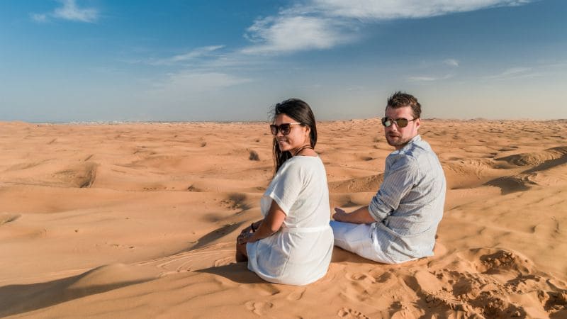 A couple seated on the smooth sands of the desert, gazing at the horizon together.