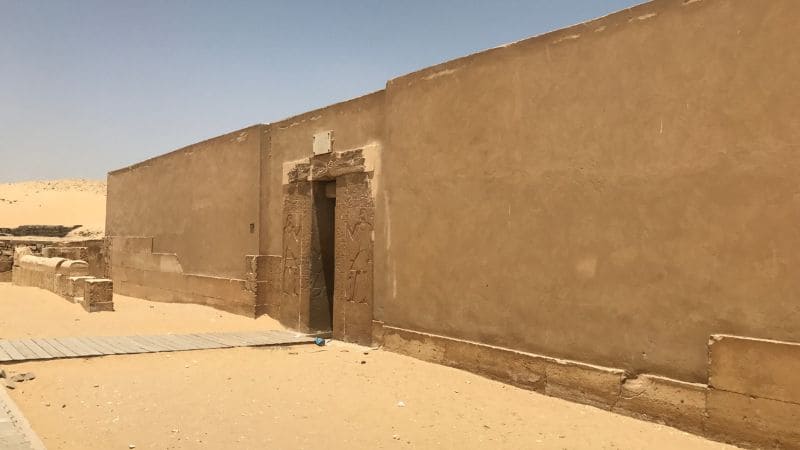 The exterior of a simple, mud-brick structure in an Egyptian desert landscape.