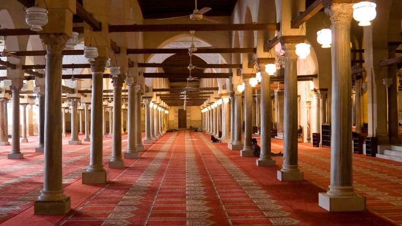 Inside a mosque, a corridor lined with columns and illuminated by hanging lamps.