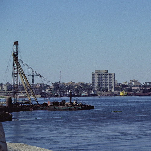 A crane on a barge in a river with a cityscape in the background under a clear blue sky