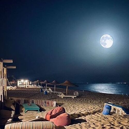 A tranquil beach at night with a large moon reflecting on the sea, beach chairs, and bean bags on the sand