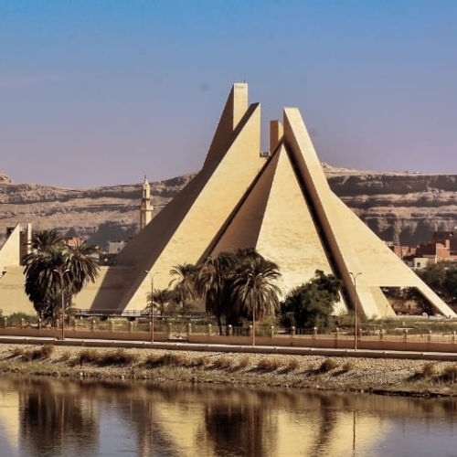 A modern, pyramid-shaped building with sharp angular design beside a palm-fringed river.