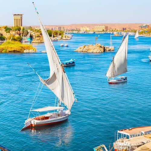 Traditional felucca sailboats on the blue waters of the Nile River with Aswan cityscape in the background.