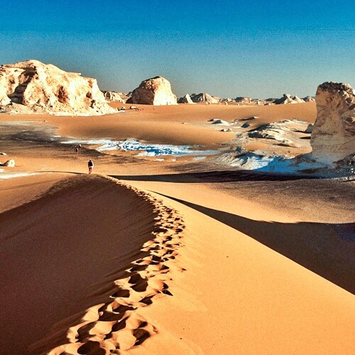 A lone hiker traversing the sculpted dunes and rock formations of a desert landscape with patches of snow.