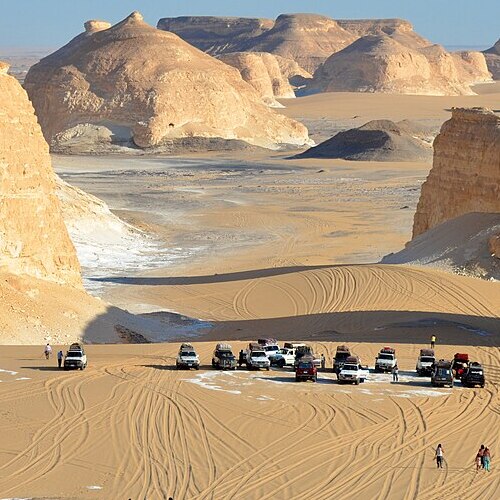 A caravan of vehicles parked in a sweeping desert landscape with towering sand formations