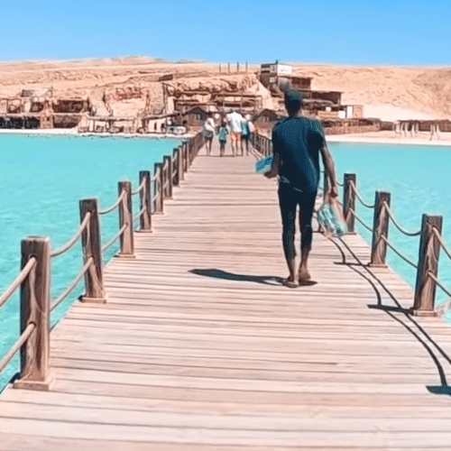 A person walking on a wooden pier at Hurghada Marina with clear turquoise waters on either side, leading to a rustic shoreline establishment under a bright blue sky