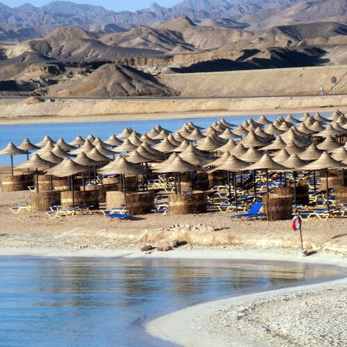 A tranquil beach scene with straw umbrellas and sun loungers overlooking a serene lake with rugged mountains in the background