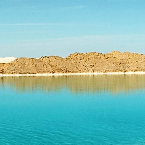 A tranquil turquoise lake with sandy hills in the background under a clear blue sky