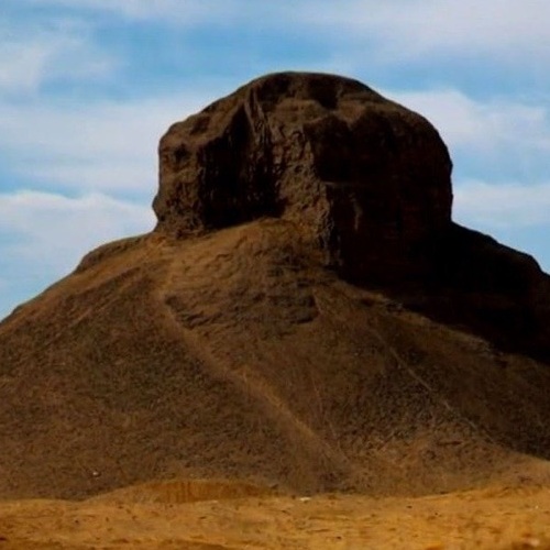An isolated hill with a striking resemblance to a human head in a desert landscape under a blue sky
