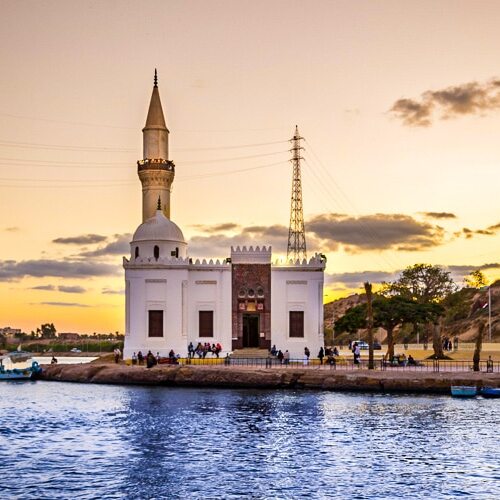 A white mosque with a golden minaret by the water's edge at sunset, with people gathered nearby