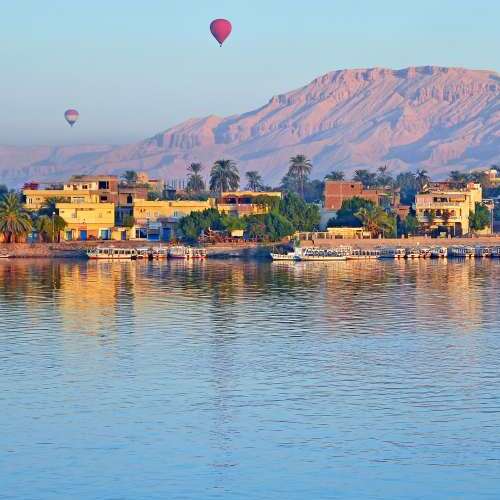 An early morning view of hot air balloons over the Nile river, with distant mountains and riverfront buildings in soft light