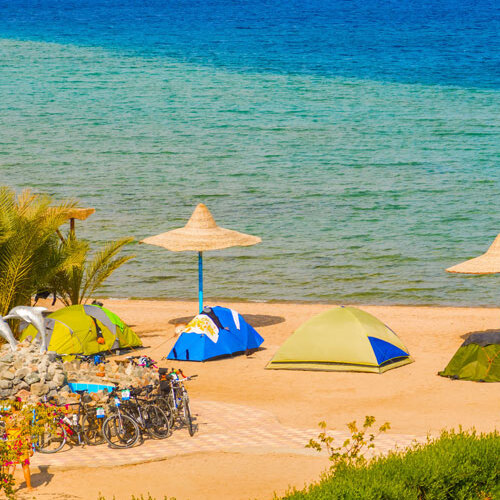 Camping tents set up on a sandy beach with straw umbrellas and bicycles, overlooking the calm blue sea