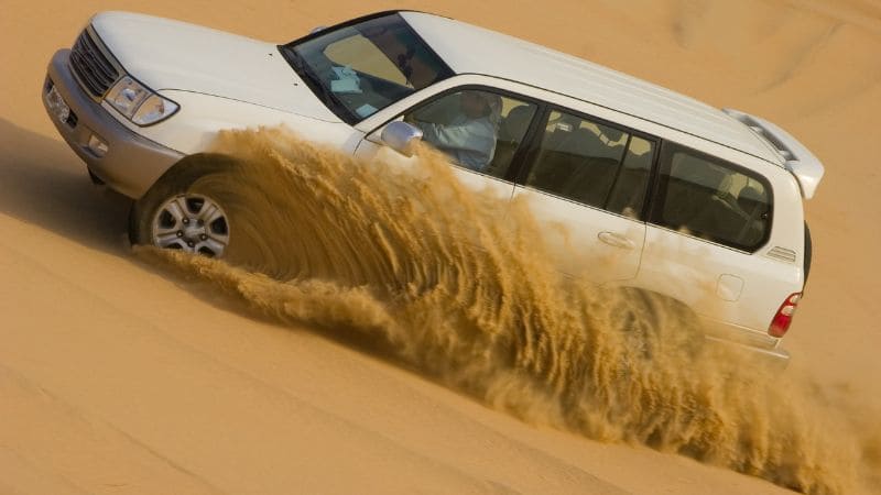 A white SUV driving swiftly through the desert, kicking up a trail of sand.