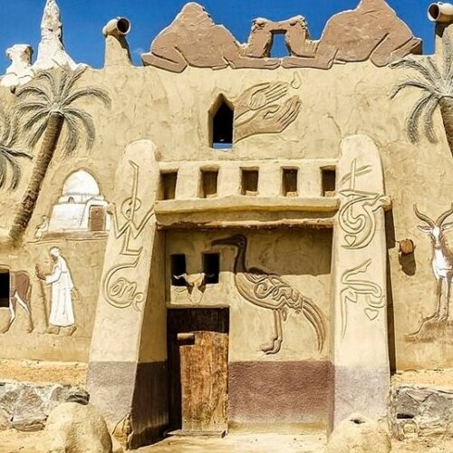 An adobe-style mud-brick fort with ornate carvings and palm trees against a bright blue sky