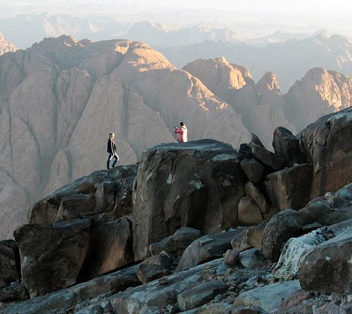 Two individuals stand on separate rocky outcrops overlooking a mountain range at dawn