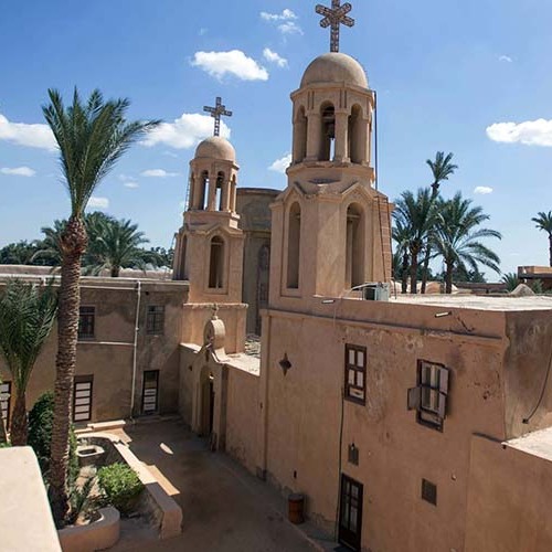 An aerial view of the Bramous Monastery in Egypt, showcasing two bell towers and a courtyard with a palm tree