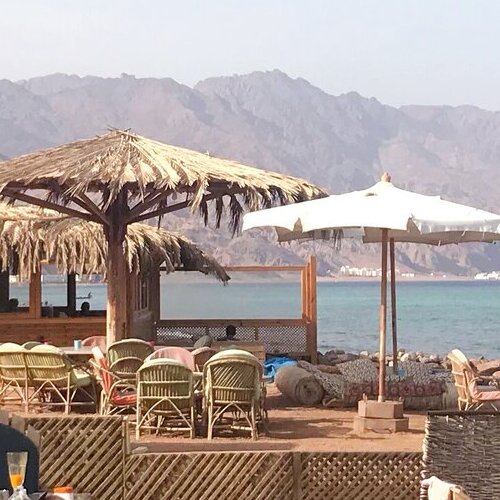 A cozy beachside setting with thatched umbrellas and wicker chairs facing the calm blue sea, with rugged mountains in the background