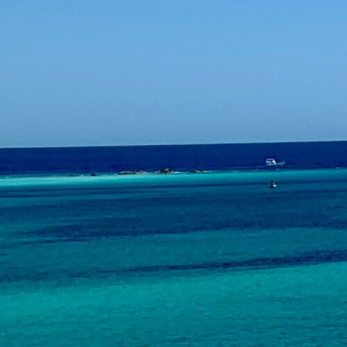 Graduated hues of blue in the sea with a distant sandy islet and a solitary boat under a clear sky