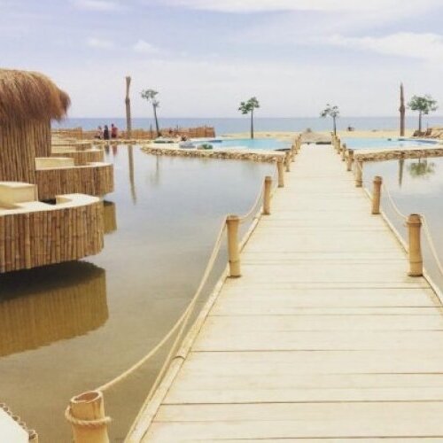 A wooden walkway leading through a tranquil resort with thatched cabanas and palm trees beside a calm water body, under a cloudy sky