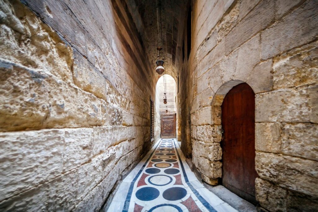 A narrow, historical alley with high stone walls, featuring a colorful, circular patterned floor and hanging lanterns leading to a wooden door at the end