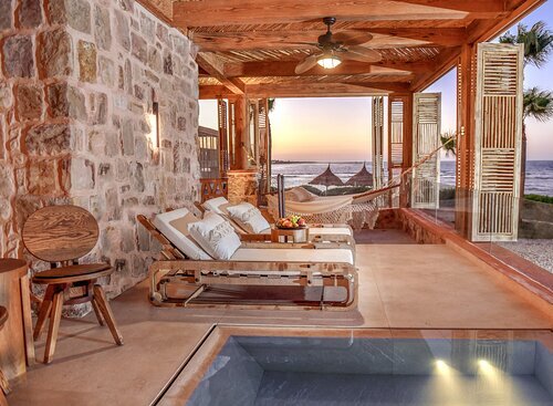 Luxury beachfront patio with stone walls, a hammock, lounge chairs, and a plunge pool overlooking the ocean at sunset