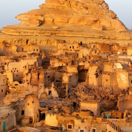 Ancient, sunlit mud-brick buildings at the base of a towering rock formation