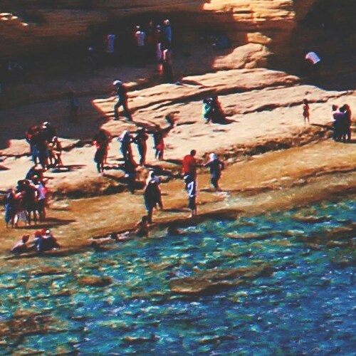 A bustling beach scene with people enjoying the sun along a rocky coastline, overlooking vibrant blue waters