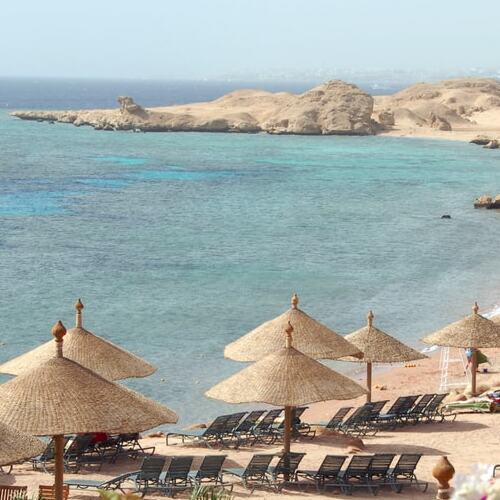 Straw parasols and loungers on a sandy beach overlooking a clear turquoise sea with rocky outcrops in the distance