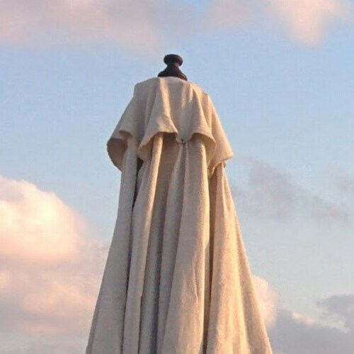 A person draped in a flowing cape stands facing a cloudy sky, exuding a sense of contemplation and serenity