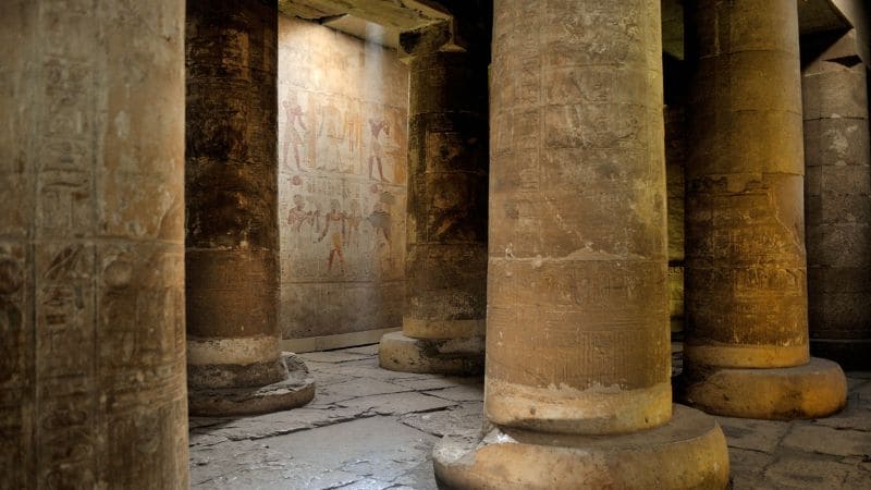 Shadowy interior of an Egyptian temple with columns and faded wall carvings.