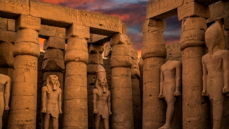 Statues and pillars within an ancient Egyptian temple complex during sunset.