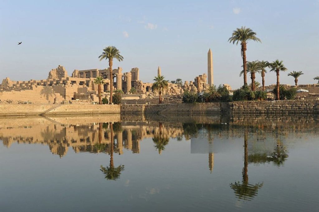 View of Karnak Temple complex reflecting in a water body with palm trees.