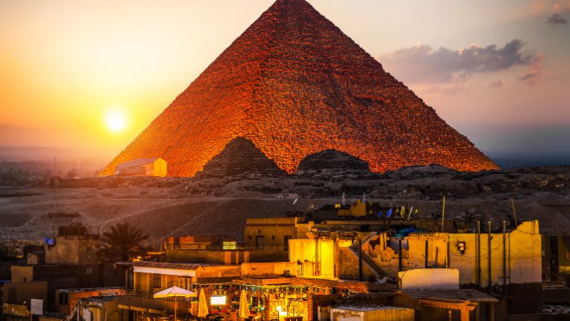 Sunset illuminating the Great Pyramid of Giza, with nearby buildings lit up in the foreground.