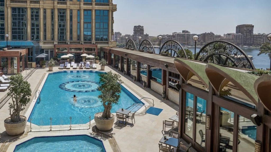 A large swimming pool at a hotel, flanked by sun loungers and shaded seating areas, overlooking a bustling cityscape and river.