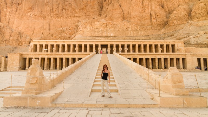 A woman stands at the centre of the wide stairway leading up to the grand Temple of Hatshepsut, carved into the rocky cliffs of Deir el-Bahari, Egypt.