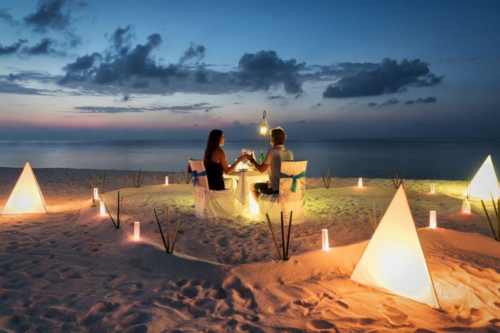 A couple enjoys a romantic dinner on the beach, surrounded by triangular lanterns and a serene ocean backdrop at sunset.