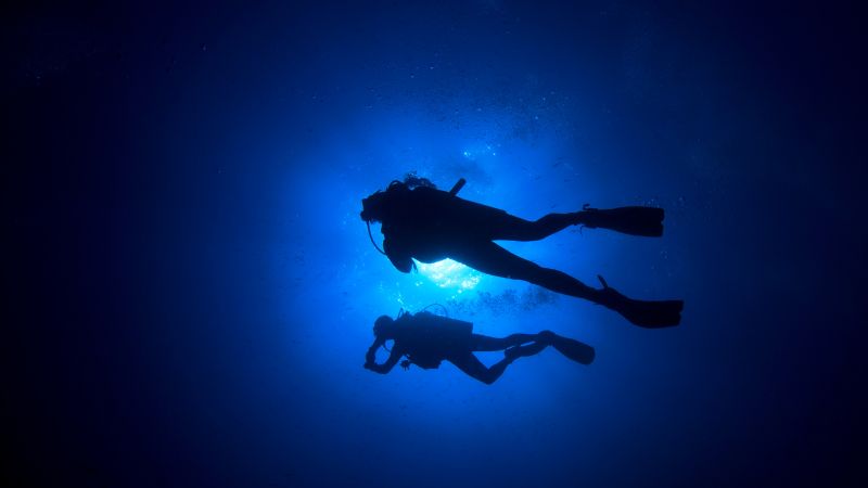 Silhouettes of two divers descend into the deep blue sea illuminated by a beam of light