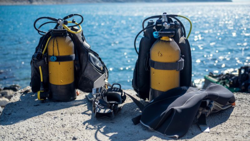 Scuba diving gear laid out on a rocky shore beside sparkling sea waters