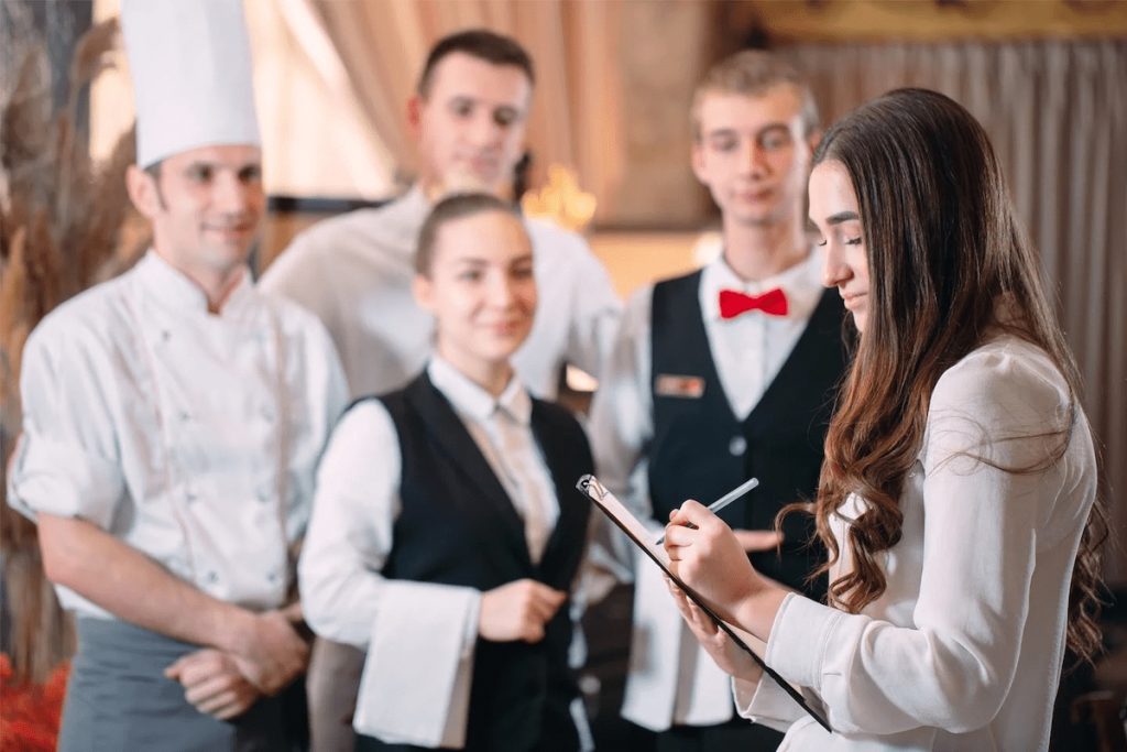 Focused young female manager writing on a clipboard with a chef in white and three waitstaff members in uniforms standing behind her in a restaurant setting.