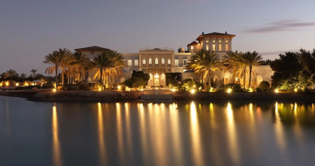 Night view of a grand estate with ornate architecture and lush palm trees, all beautifully lit and reflected in the calm waters in front.